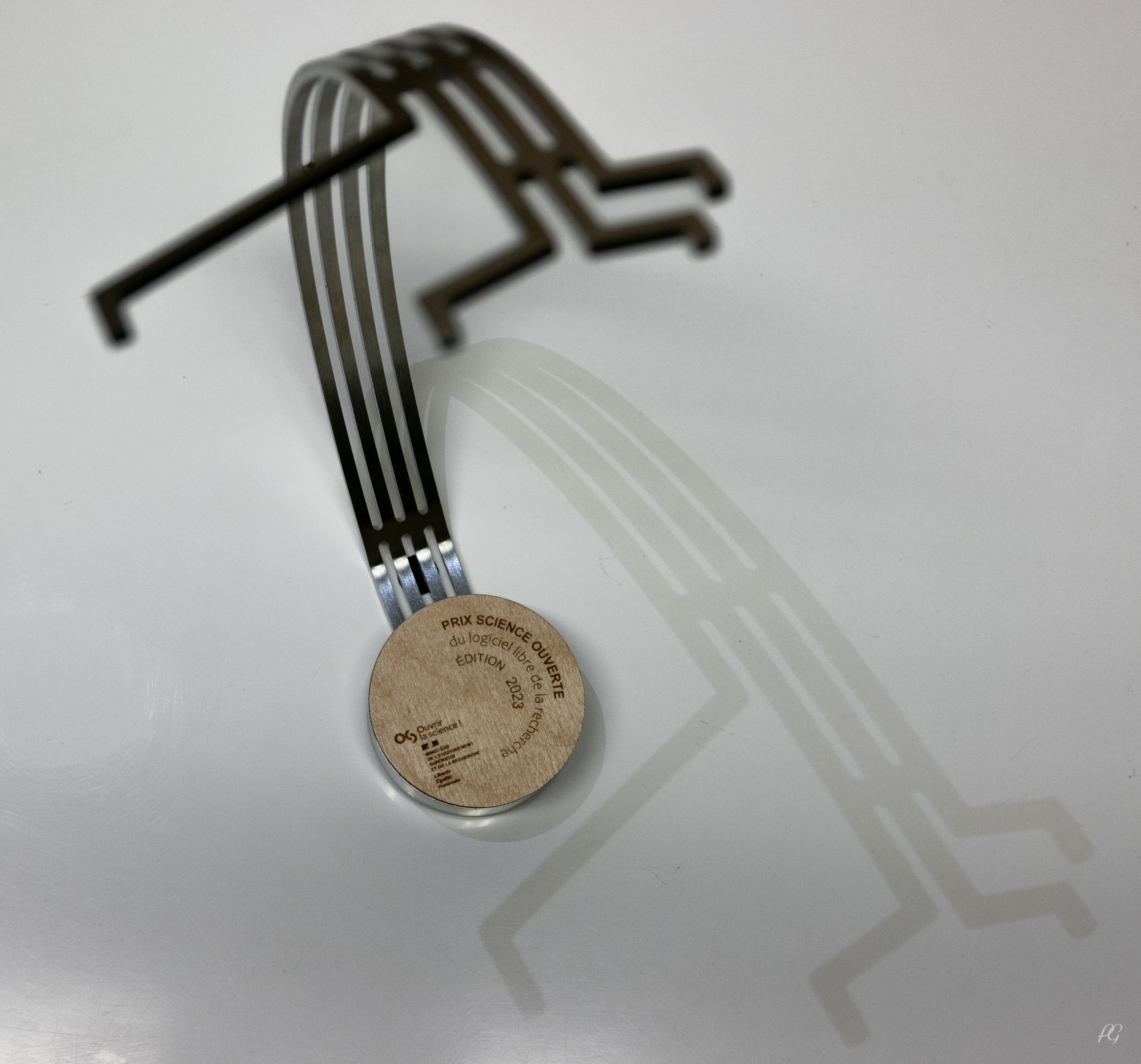 Trophy of the Open Science price seen from a above; trophy in metal with round wodden plaque at the bottom, stating 'Prix science ouverte du logiciel libre de la recherche. Édition 2023'. Four metal bars curve upwards, branching out non-symmetrically.